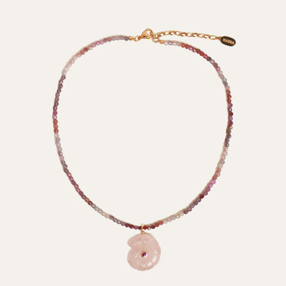 A delicate beaded necklace with a pink stone pendant, featuring a gold chain and clasp with a small brand tag that reads 'KAOUA'. The beads transition from pink and purple to clear and white, with a central pink stone shell pendant containing a tiny red gemstone.