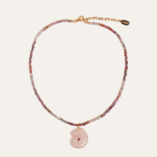 A delicate beaded necklace with a pink stone pendant, featuring a gold chain and clasp with a small brand tag that reads 'KAOUA'. The beads transition from pink and purple to clear and white, with a central pink stone shell pendant containing a tiny red gemstone.