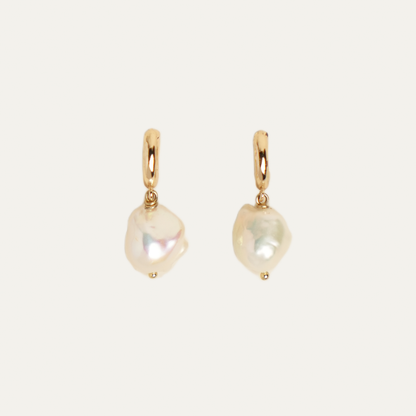 A pair of gold hoop earrings with irregular, baroque-style white freshwater pearls dangling from them, set against a solid off-white background.